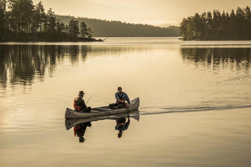 Zwei Männer fahren in einem Kanu auf einem See. Einer der beiden paddelt, der andere hält eine Angel in den den Händen. Das Licht des Sonnenaufgangs taucht die Szene in warmes Licht.