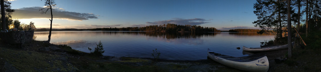 Im Vordergrund sind Kanus neben Kiefern am Ufer einer Insel zu sehen. Im Hintergrund geht die Sonne über einem See in Südschweden unter. Am Horizont sind weitere Inseln zu sehen.